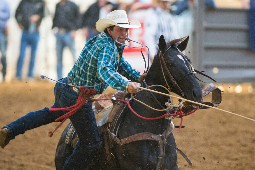 An image of Haven Meged jumping off a black horse with a rope in his hand. He is wearing a green plaid shirt, dark denim jeans, and a straw cowboy hat.