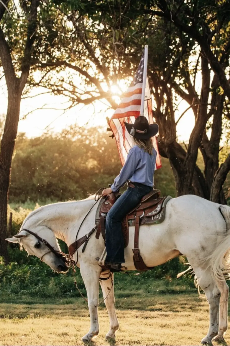 A girl wearing a black felt cowboy hat sits atop a white horse. She holds an American flag, and the sun is setting behind her. 